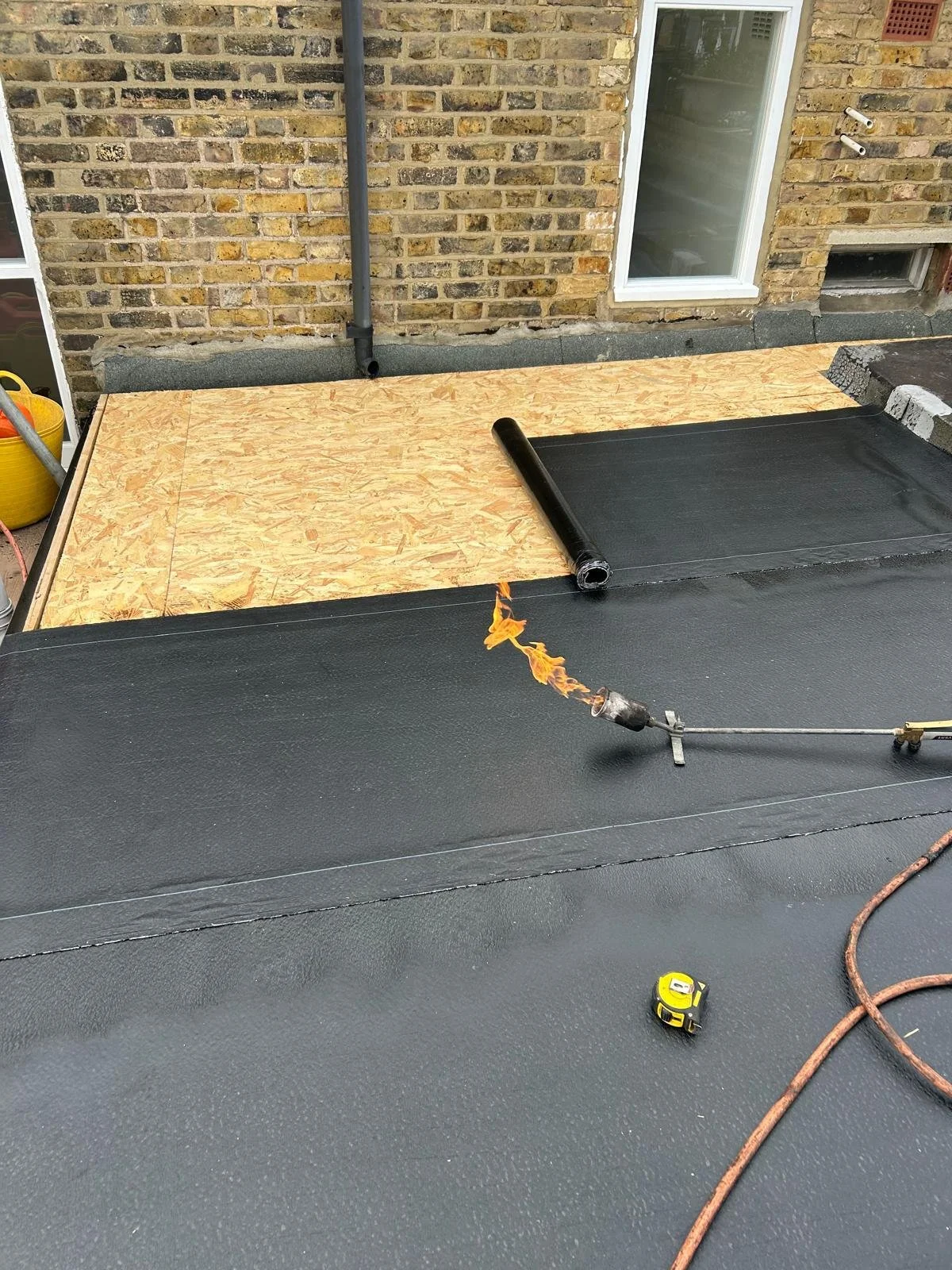 Roof under construction with black waterproof membrane being unrolled over plywood sheathing, with a propane torch heating the material.