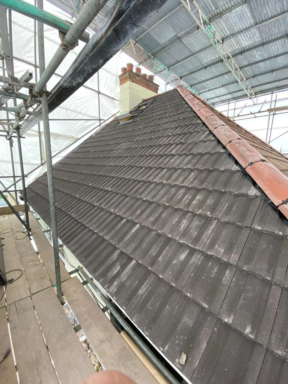 A sloped rooftop with dark gray shingles, a red ridge cap, and two small skylights, surrounded by construction scaffolding and a protective covering.