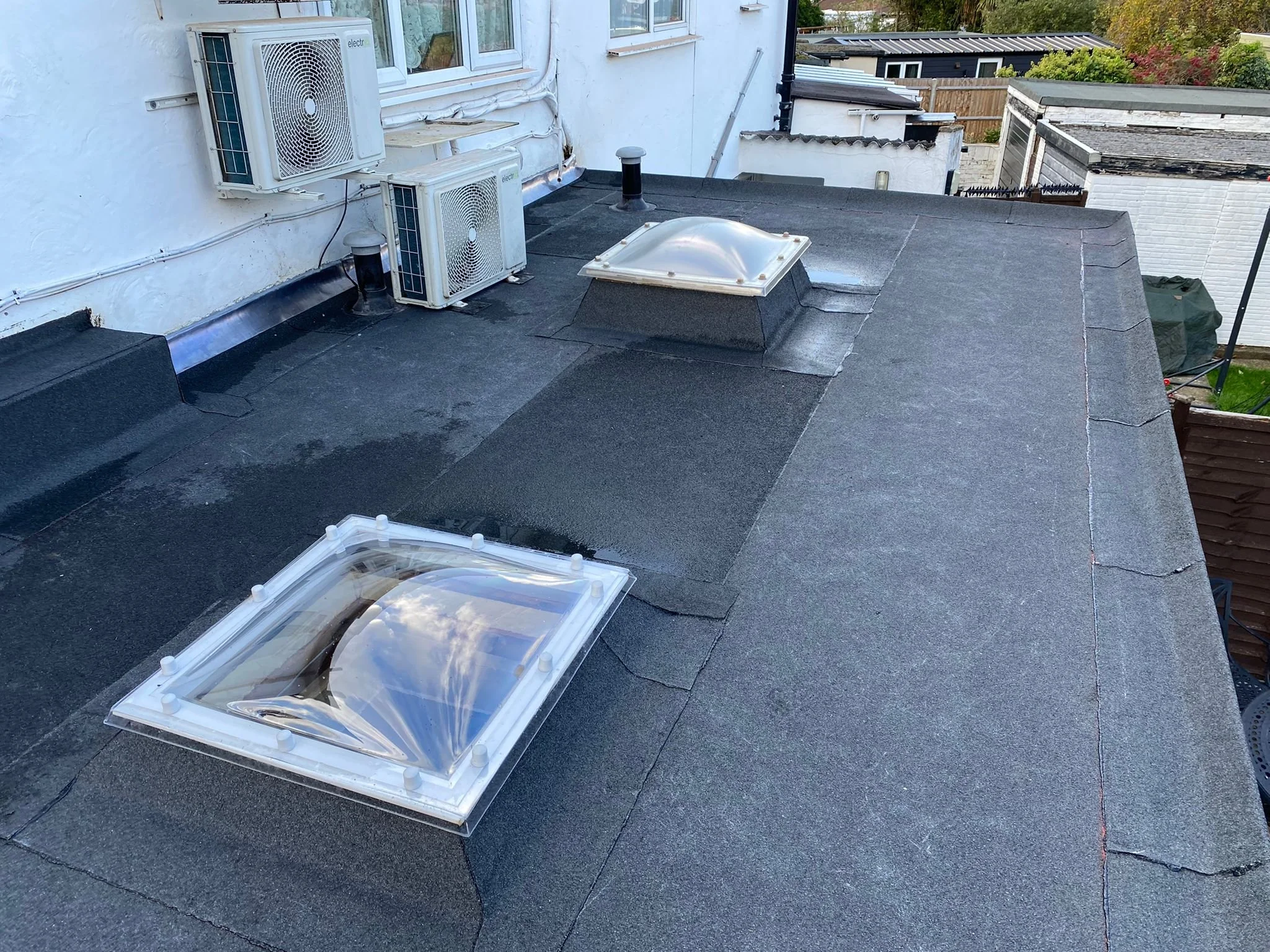 View of a flat black roofing surface with two skylights and three air conditioning units on the side of a building, with trees and rooftops in the background.