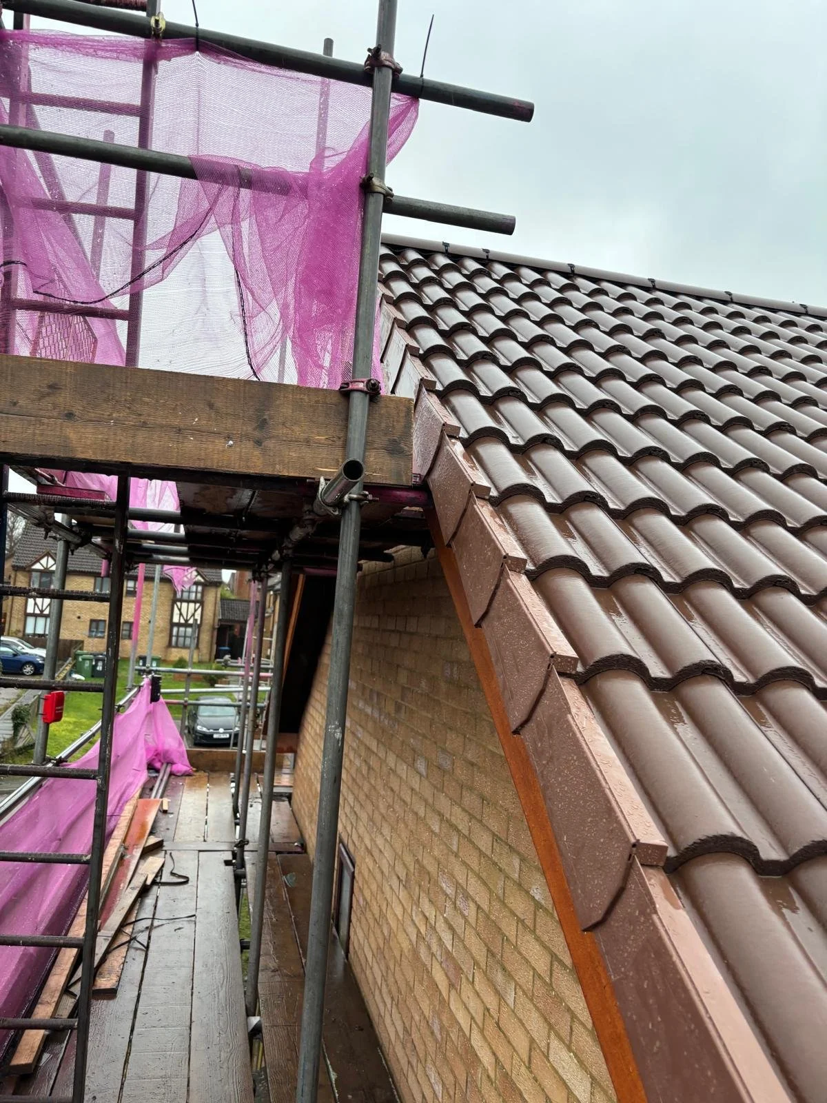 Construction scaffolding with pink safety netting next to a sloped roof with brown tiles on a brick building.
