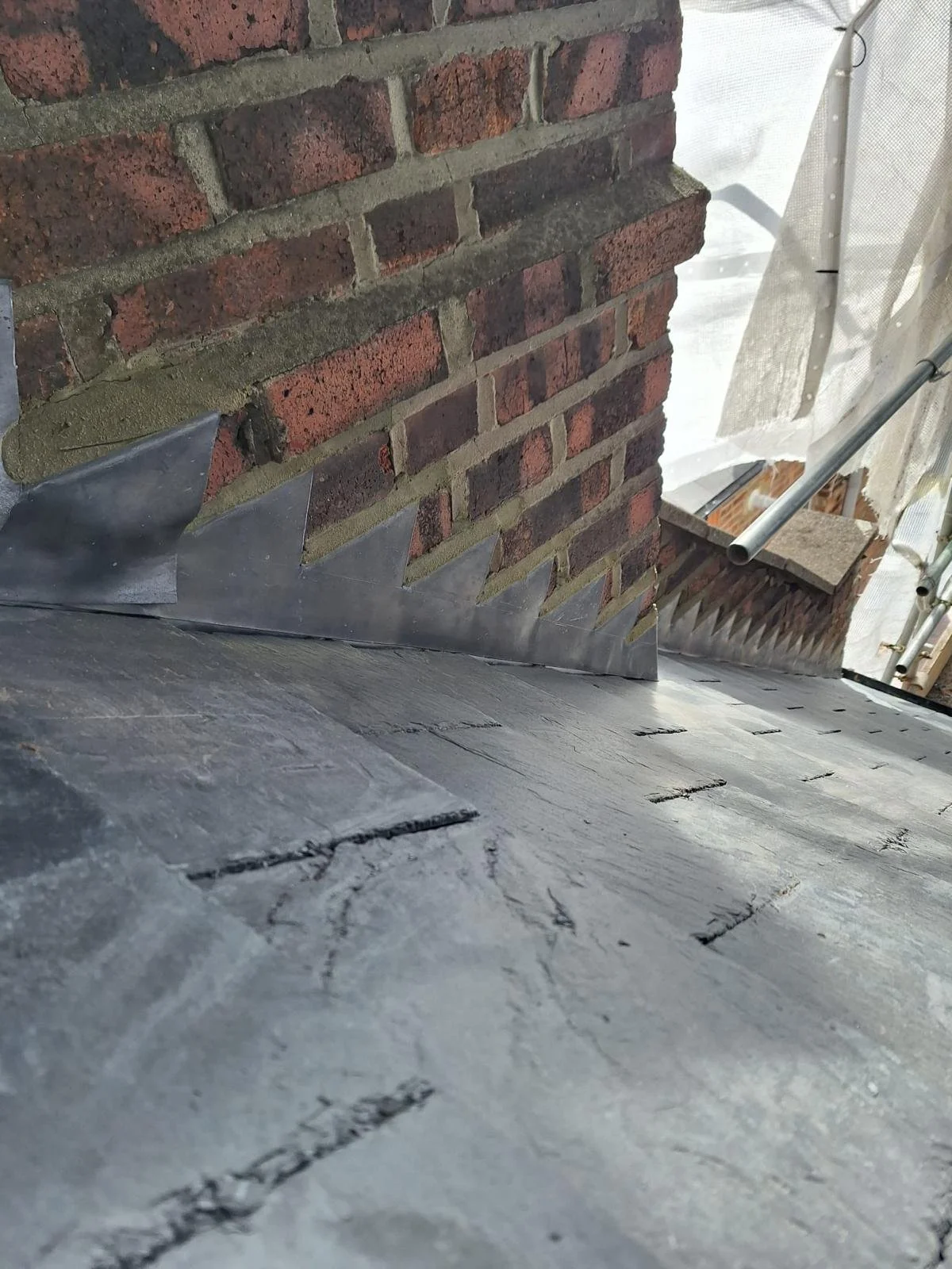 Close-up of a brick wall with a metal flashing installed at the base, and scaffolding with a metal pipe in the background.