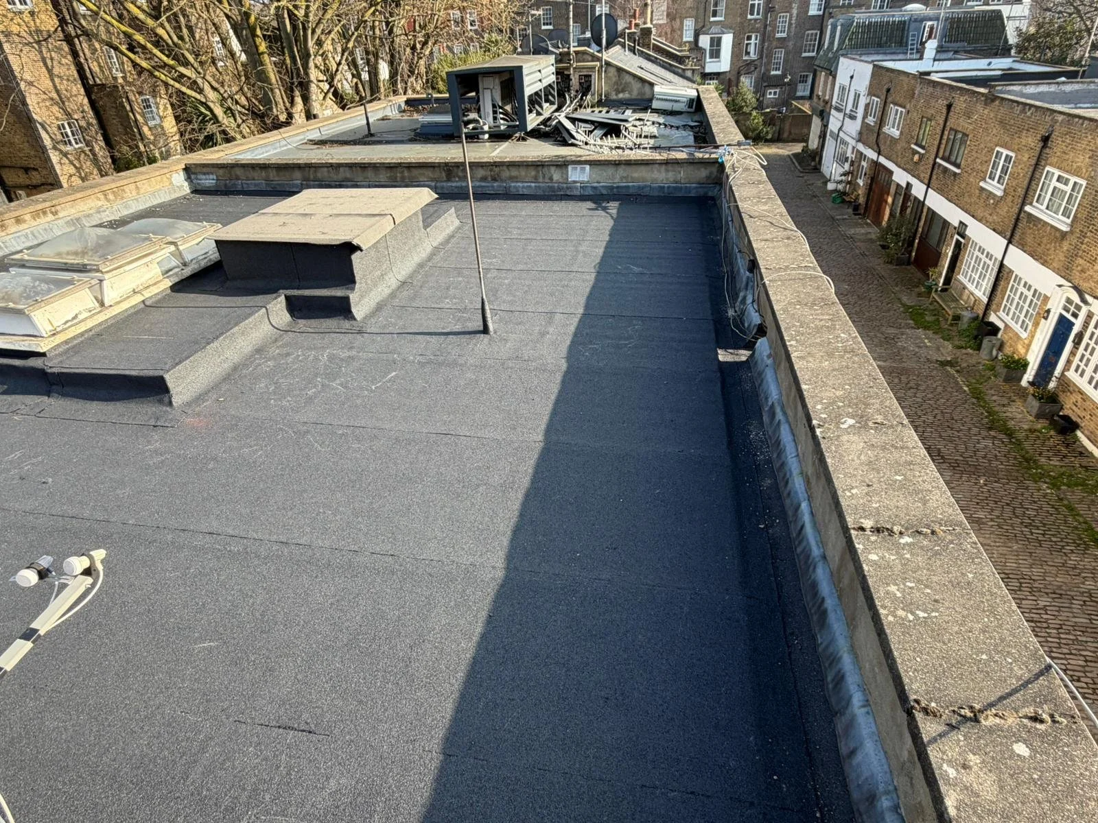 View of a flat rooftop with black waterproofing, a chimney, skylights, and a vent pipe, with neighboring secured residential buildings and a cobblestone street below.