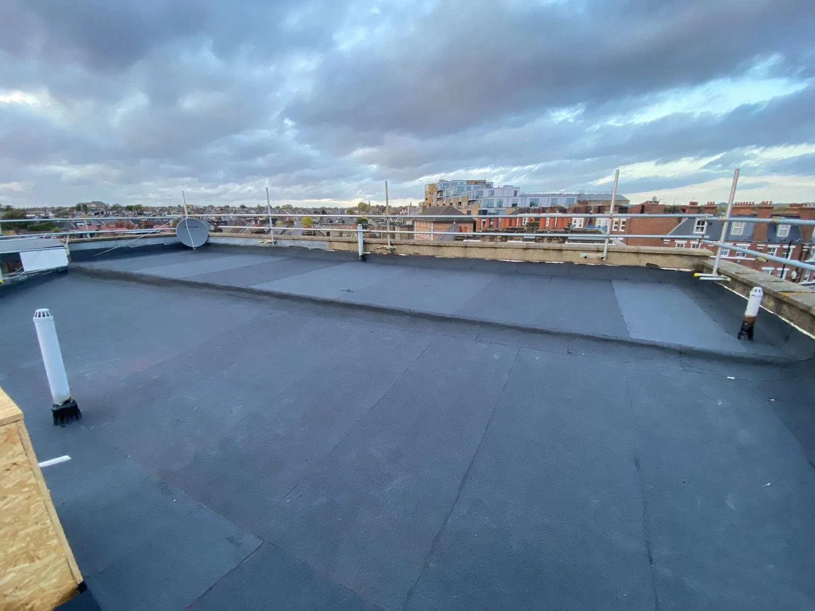 View of a flat rooftop with black waterproofing, skylights, and safety rails, overlooking a cityscape with brick and modern buildings under a cloudy sky.