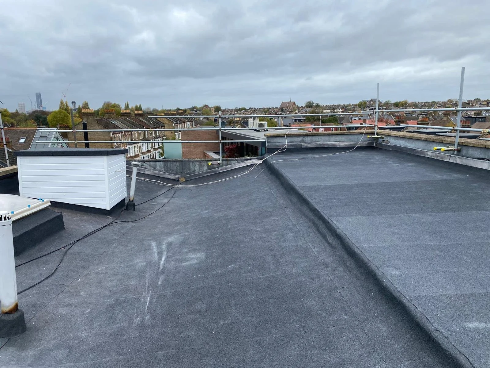 View of a building rooftop with black waterproofing material, skylights, and safety railings, overlooking a neighborhood with residential houses and cloudy sky.