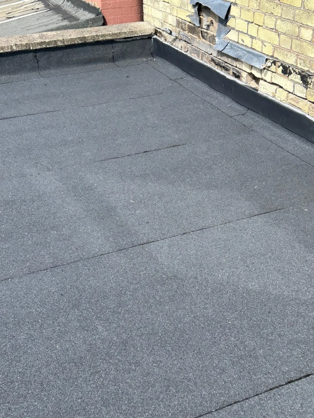 A rooftop with black gravel surface, brick wall, damaged brickwork, and metal flashing along the edge.