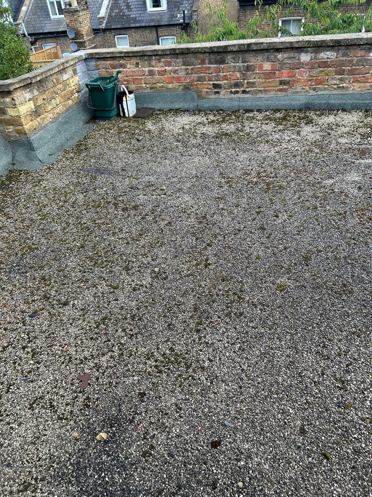 A rooftop area with gravel, a brick wall, and some garden tools including green watering cans and a white container. In the background are neighboring buildings with brick walls and windows.