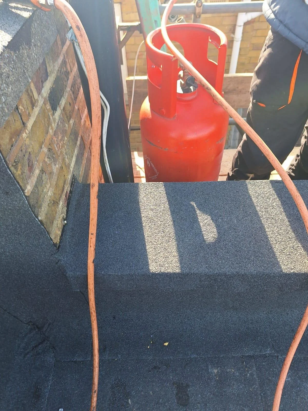 Close-up of a red propane tank on a rooftop with black roofing material, a brick wall, and an orange hose connected to the tank.