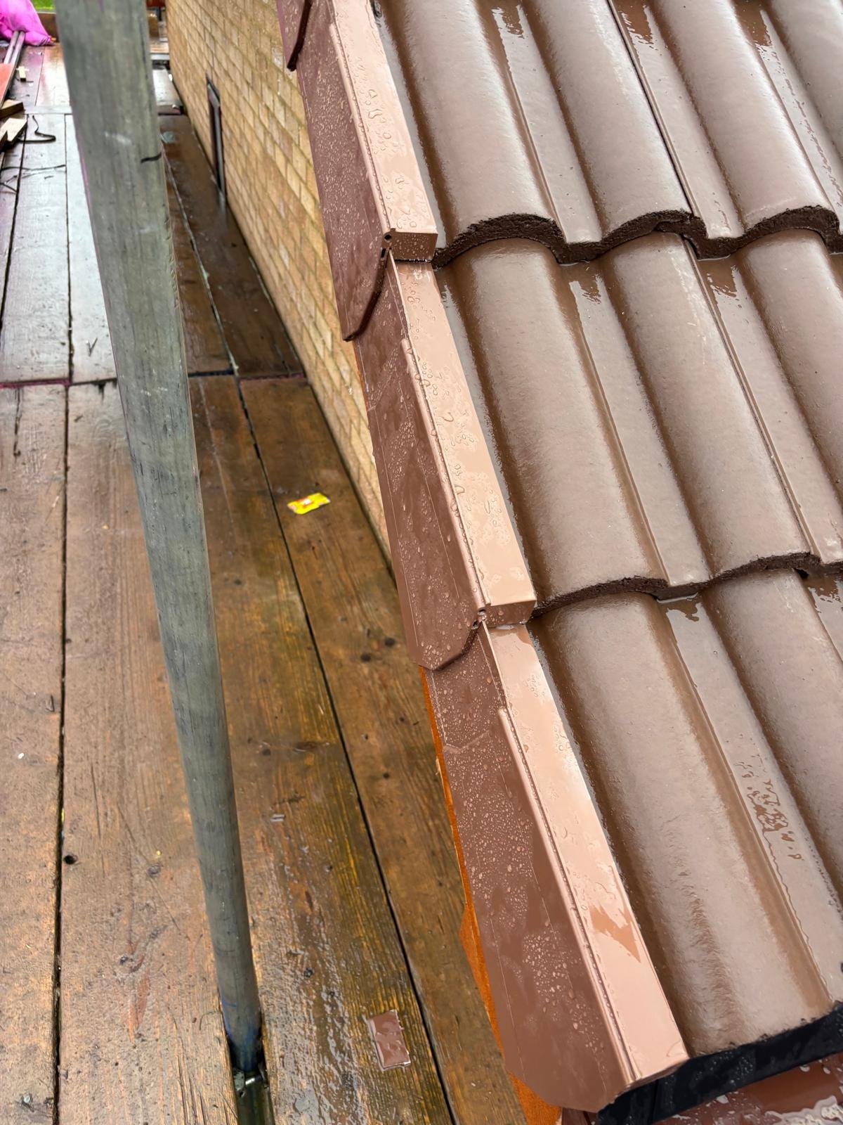 Close-up of a brown tiled roof edge with a metal drip edge flashing, next to a brick wall, on a wooden scaffold with wet wood planks.
