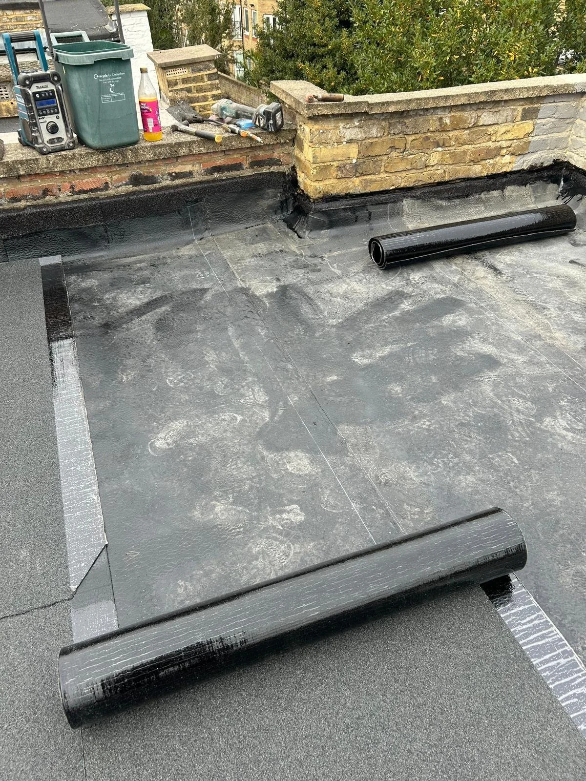 Roof surface with black waterproofing membrane, tools, and supplies on the edge, indicating roofing work in progress.