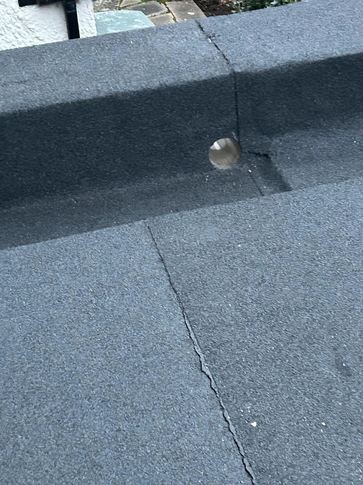Close-up of a cracked black asphalt surface with a hole in the curb, possibly for a bolt or pipe, in an outdoor setting.