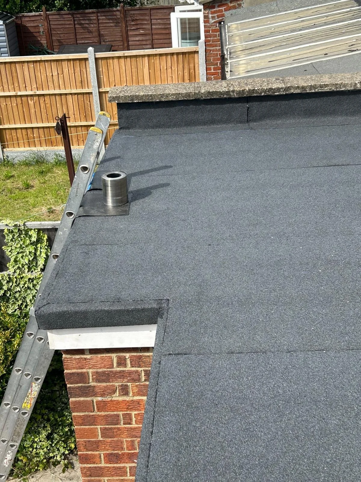 Roof with black asphalt shingles and a chimney, with a ladder leaning against the roof and a coil of roofing material on top.