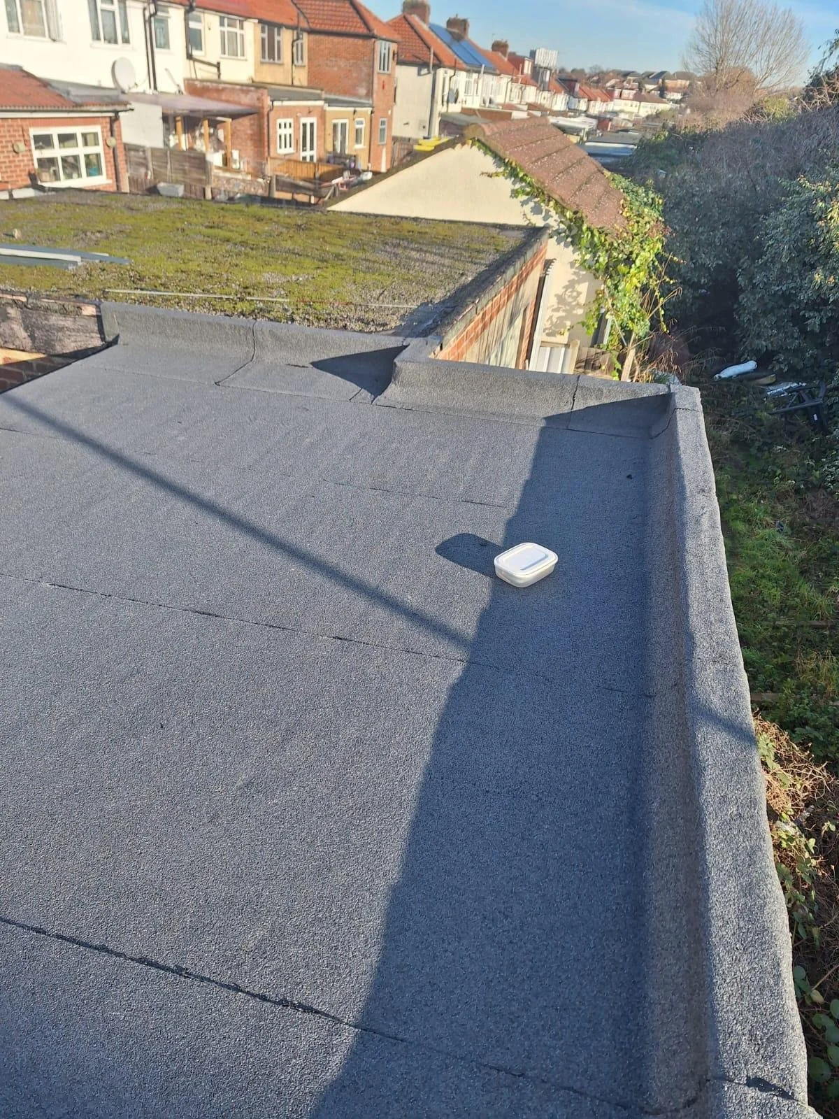 View of residential rooftops with moss-covered roof, a flat roof with a small white container, and neighboring houses with various roof styles under a clear blue sky.