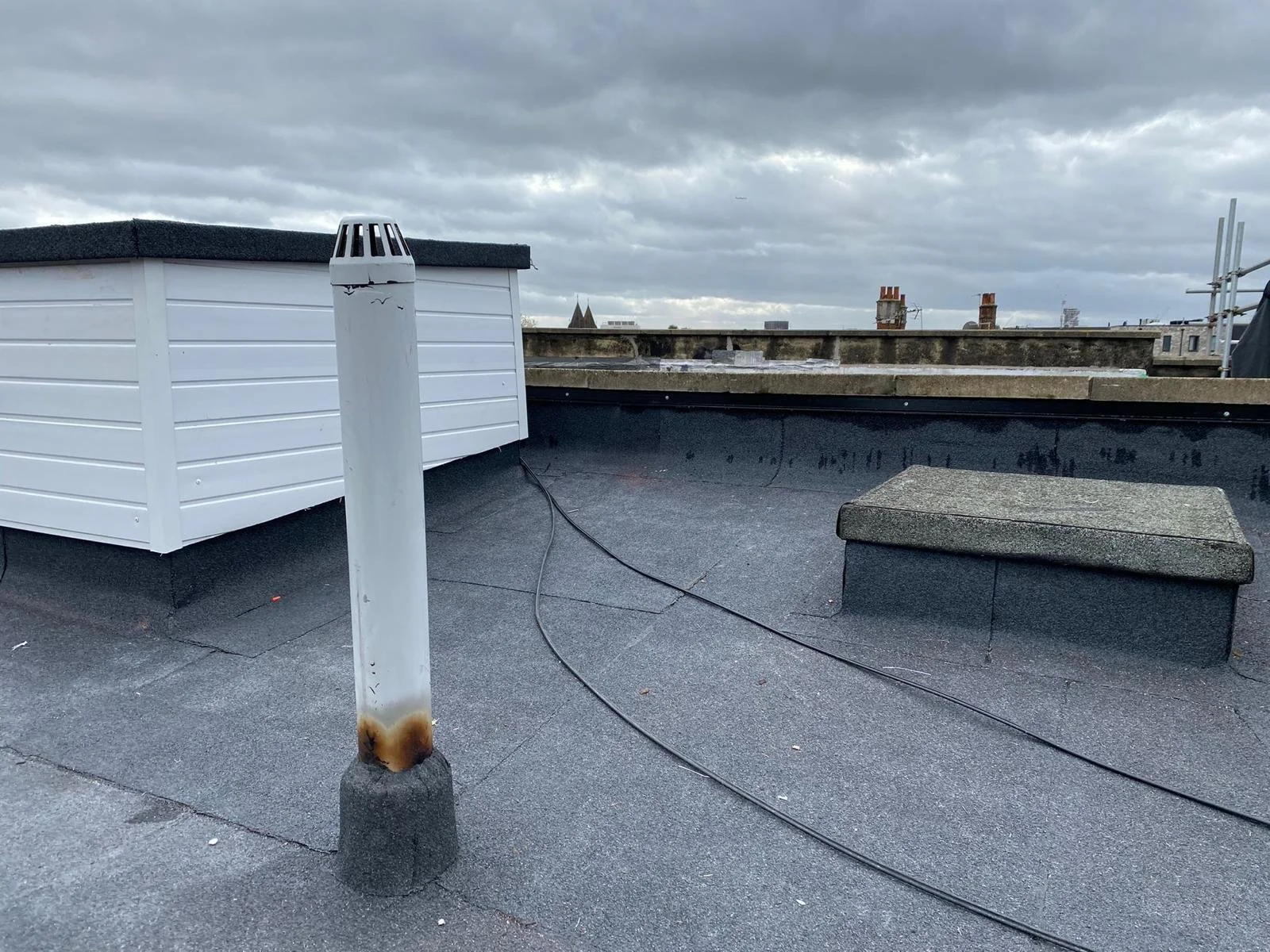Rooftop with a white vent pipe, a white utility box, and a small concrete storage bench under a cloudy sky.