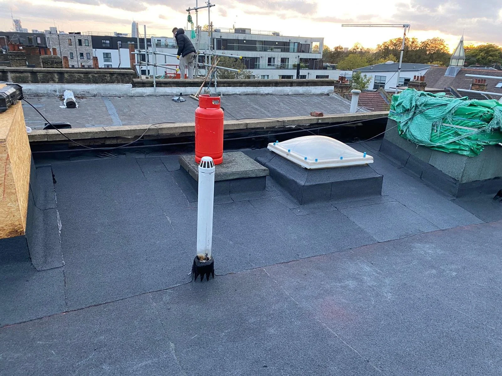 Flat rooftop with vents, a red propane tank, and construction materials, with a worker working in the background at sunset.