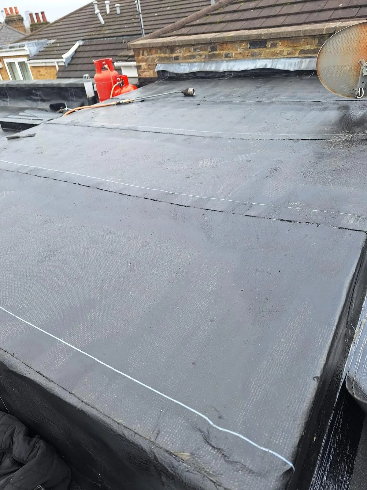 Flat roof with a black rubber membrane, two red gas cylinders, a small flashlight, and a satellite dish on the right side, with neighboring rooftops in the background.