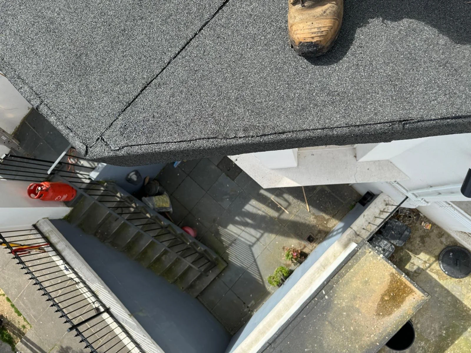 A view from a high angle showing a rooftop and a small outdoor patio. The rooftop has black shingles with a slight crack, and the patio has black tiles with some gardening items and steps leading down. A red fire extinguisher is mounted on the patio 