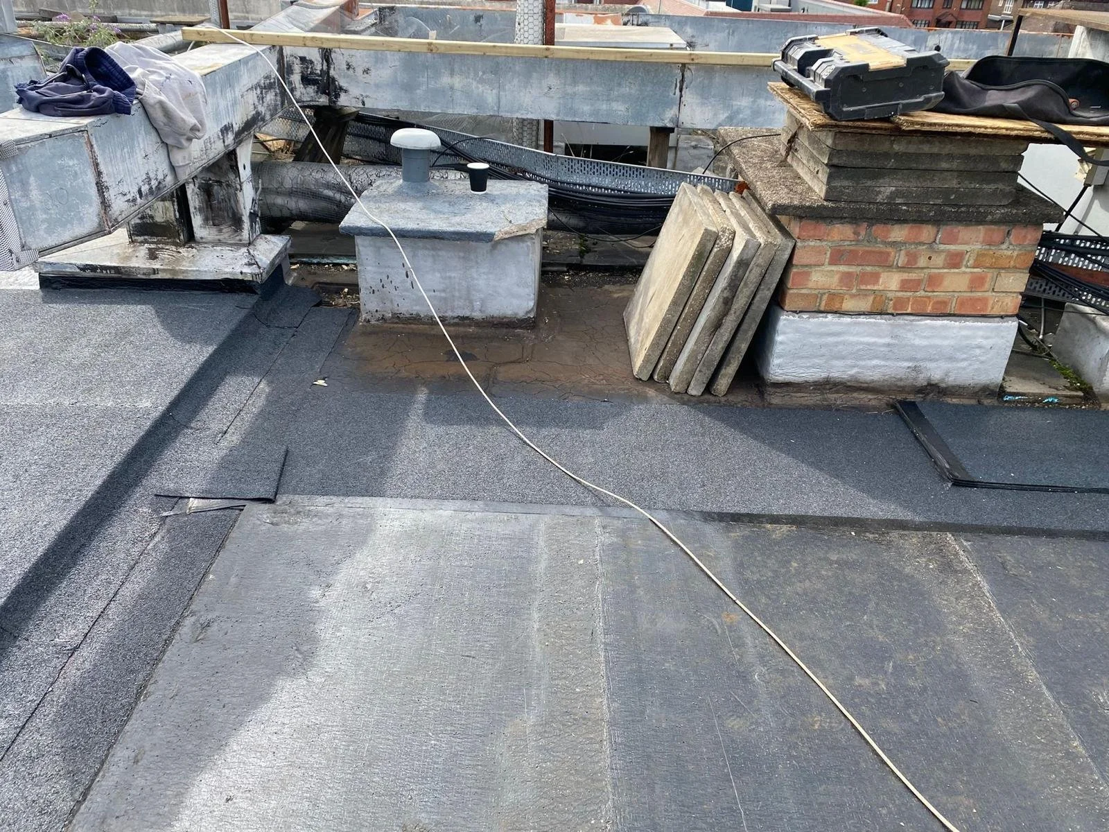 Rooftop with construction materials, black toolbox, and clothing, showing a partially wet dark gray rubber or asphalt roofing surface.
