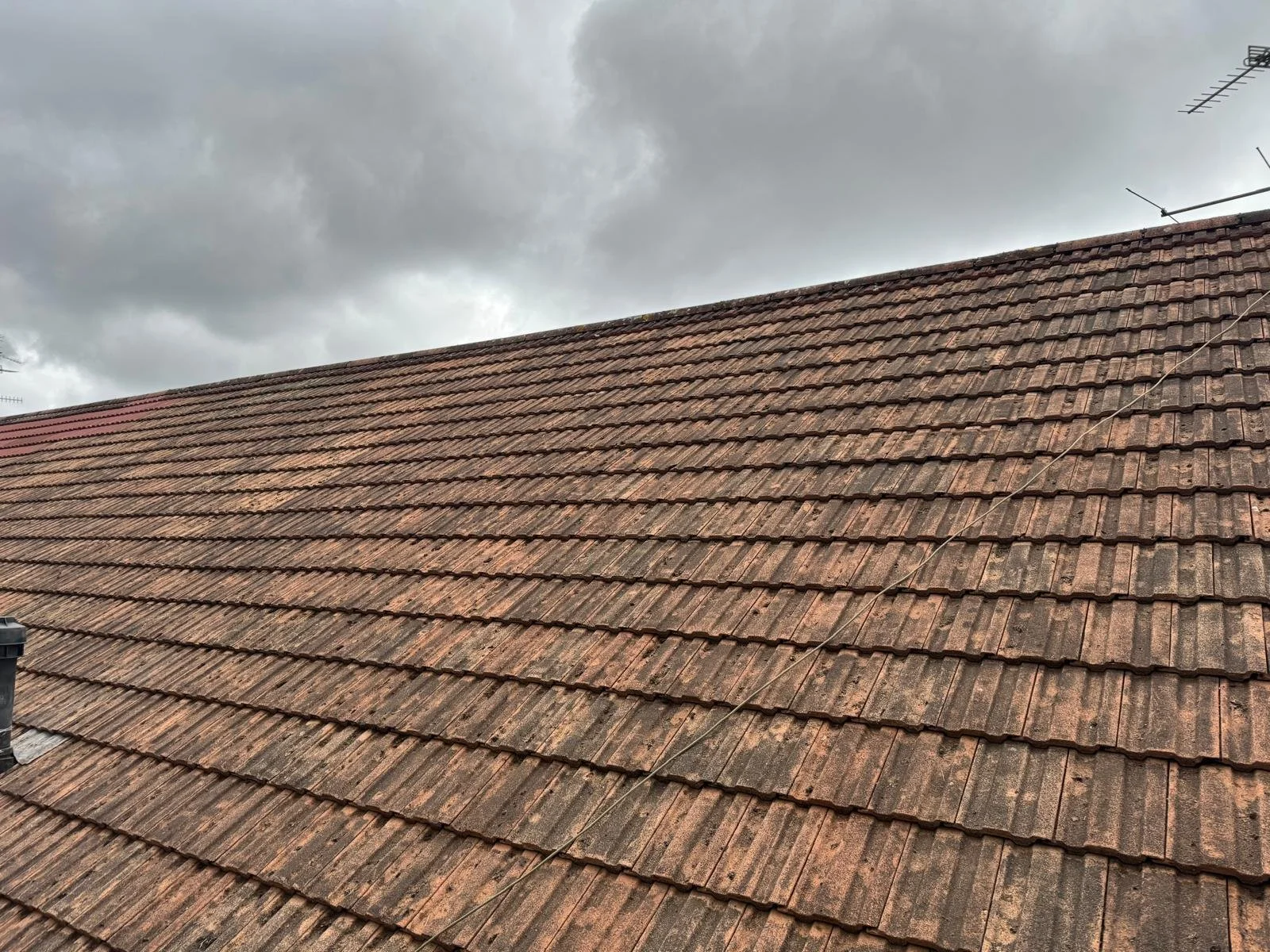Close-up view of a clay tile roof on a house under a cloudy sky, with an antenna and some wires visible.