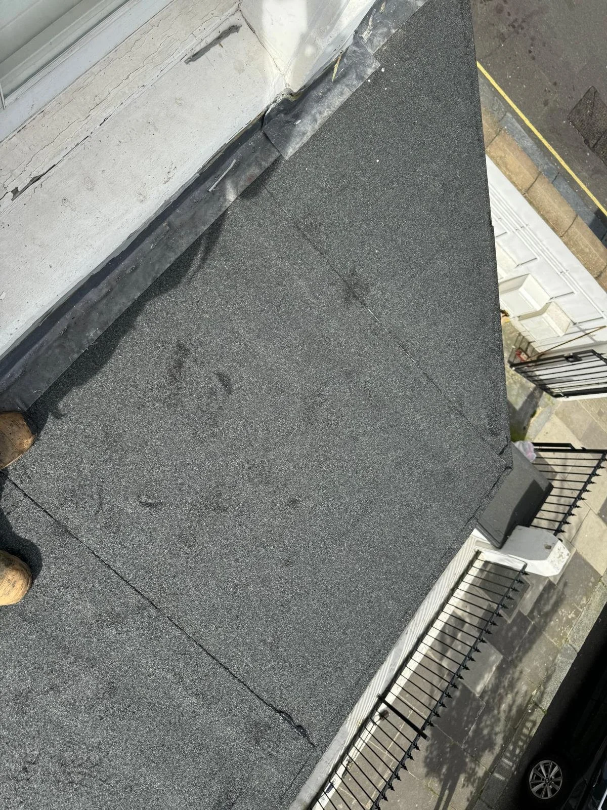 Close-up view of a flat black rubber roofing surface on a building with a white wall and black metal railing. The roof shows seams and slight wear.