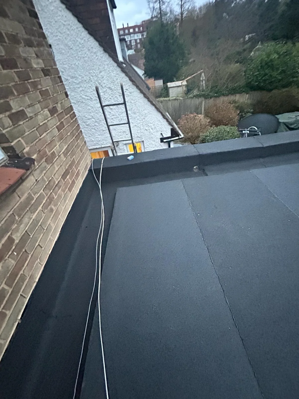 View of a residential roof with black roofing material, a brick wall on the left, and a neighboring house with a ladder against the wall. In the background, there are trees, bushes, a fence, and a barbecue grill covered with a tarp.