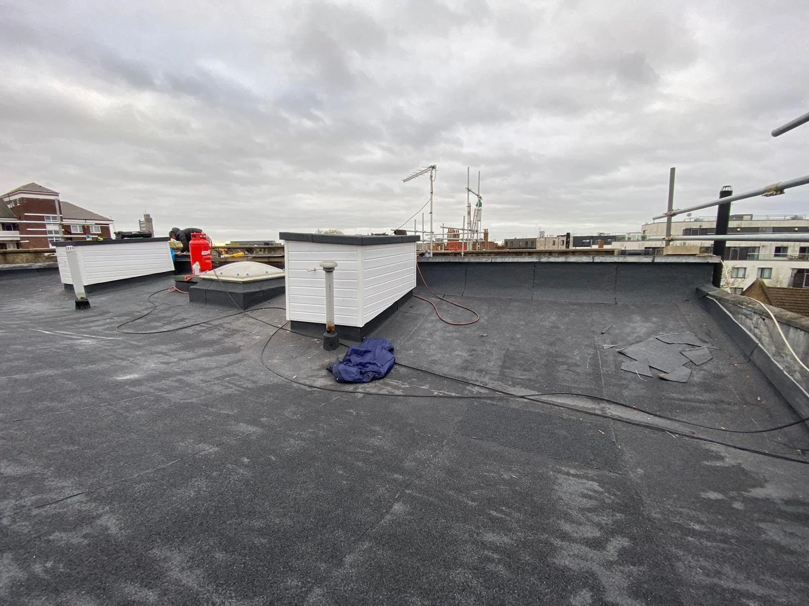 View of a rooftop with black roofing material, white rooftop structures, antennas, and a worker in the background. The sky is overcast.