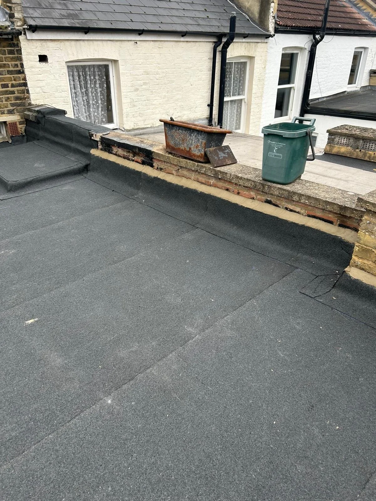 View of a rooftop with black roofing material, partially removed brick wall, and a rusty metal container; against a white brick building with three windows and black downspouts.