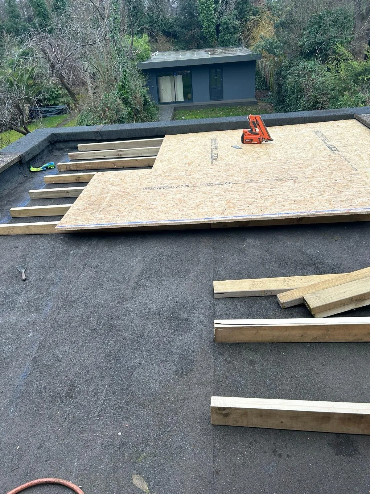 Construction site on rooftop with plywood sheets and wooden beams, a power saw, and a wrench, overlooking a backyard with trees and a small building.