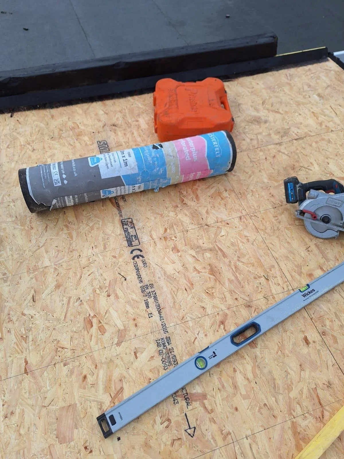 Construction site with plywood platform, level tool, circular saw, safety box, and insulation roll.