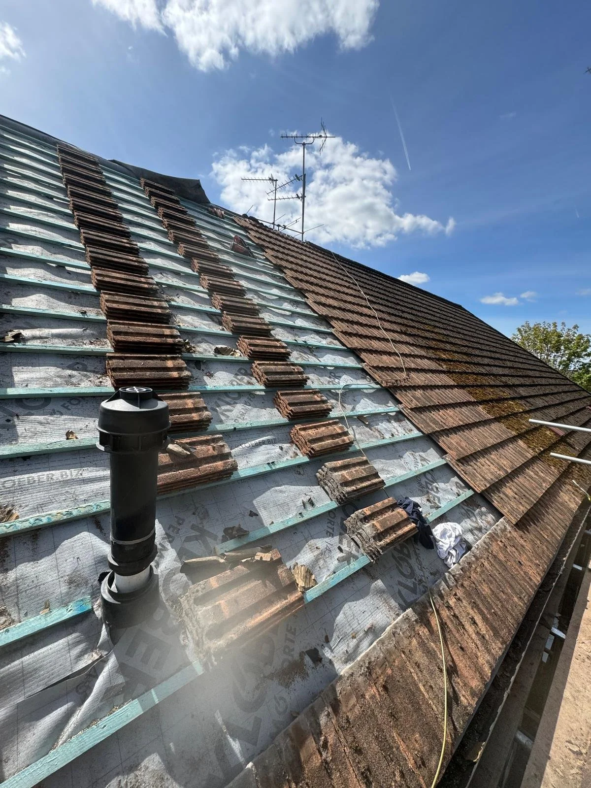Roof under repair with some tiles removed, exposing insulation and underlayment, with roofing tools and materials present, under a blue sky with clouds.
