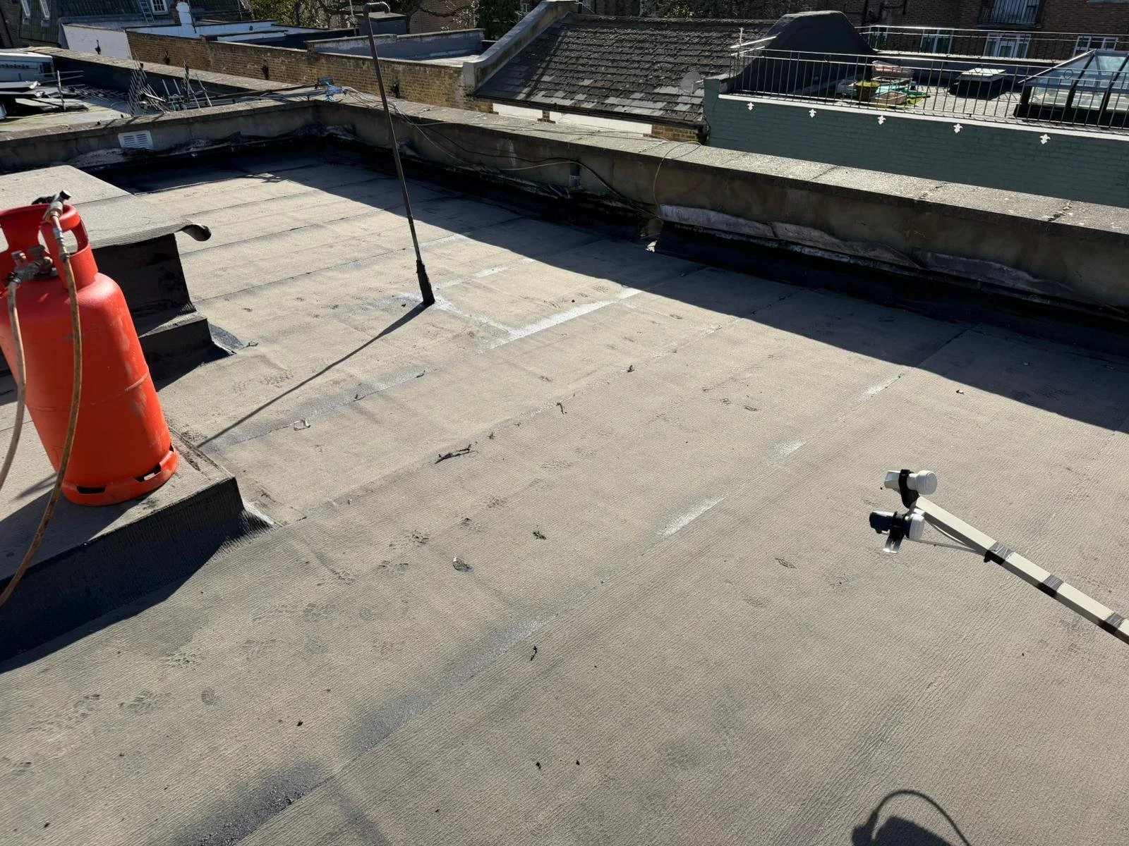 View of a flat rooftop under construction or repair, showing a red safety cone, a vertical pole, and a mounted directional device, with surrounding rooftops and building structures in the background.
