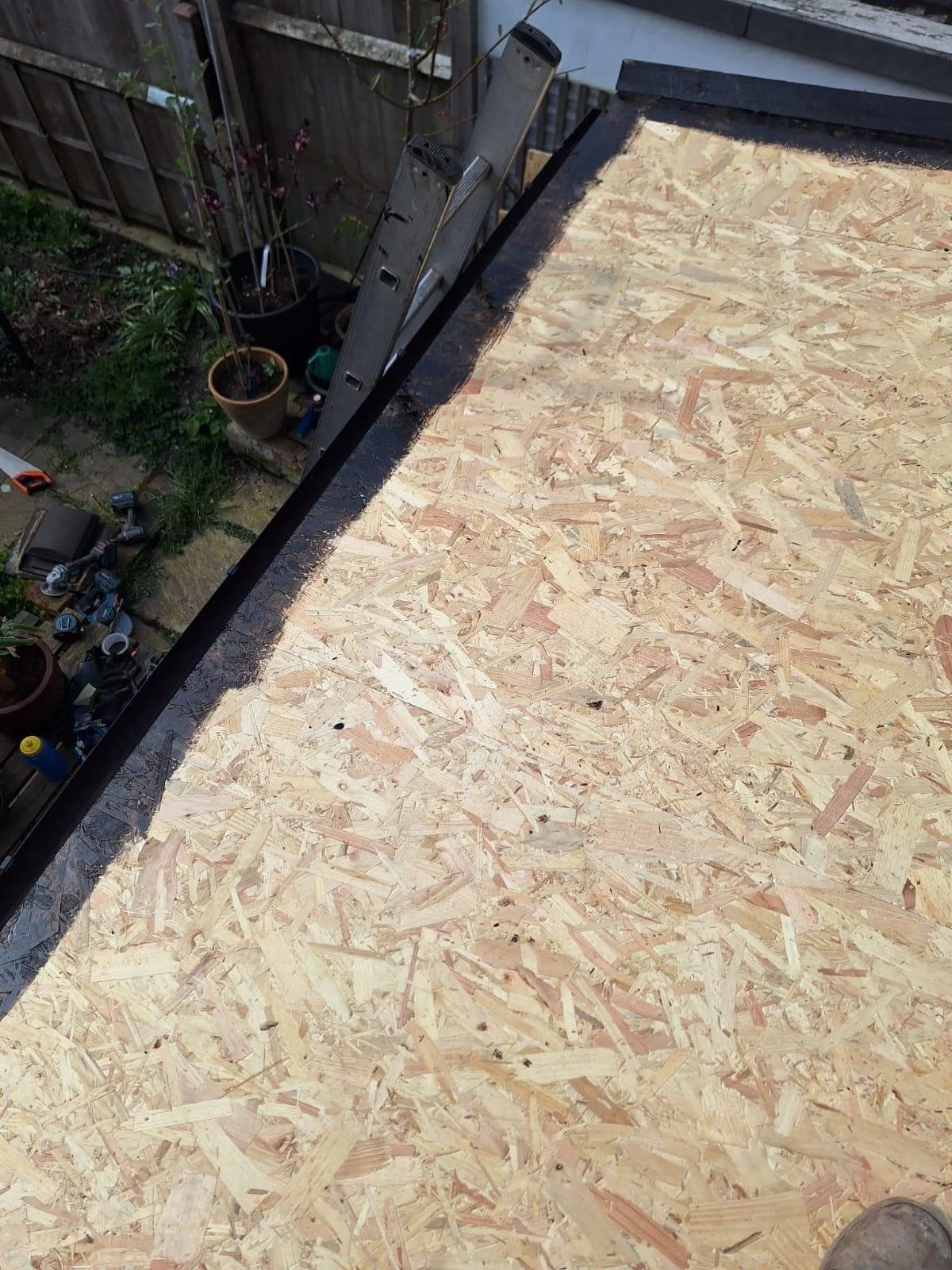 Unfinished plywood roof deck with tools and a ladder, overlooking a garden with potted plants. 