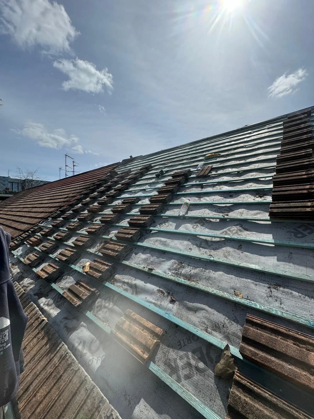 Roof under renovation with tiles and underlayment still exposed, partially covered by new tiles, under a bright sunny sky.