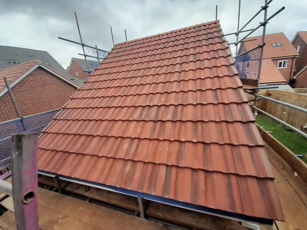A newly tiled red roof on a building under construction, surrounded by scaffolding, in a residential neighborhood with other houses nearby.