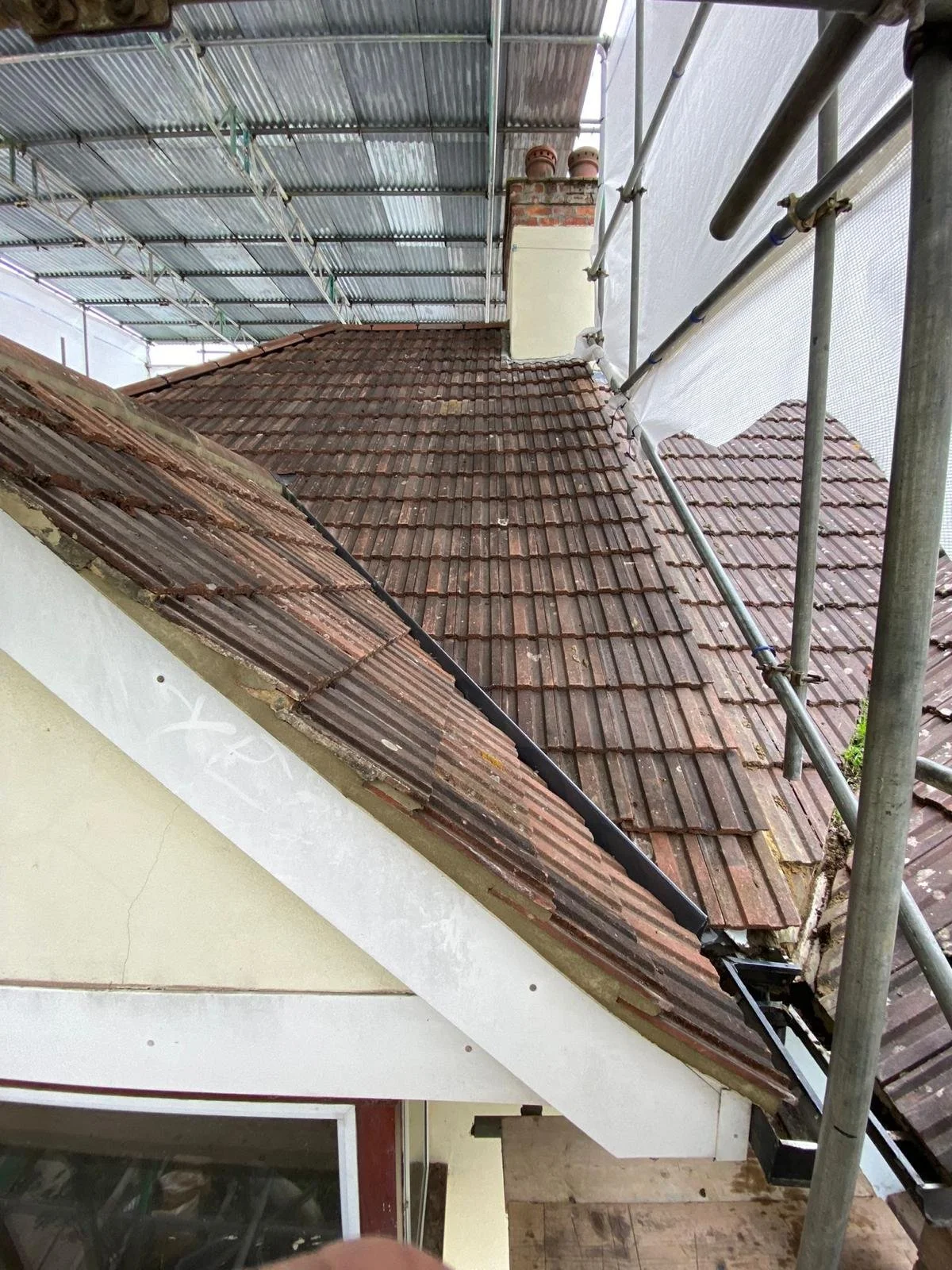 Photo showing a brown tiled roof of a house under repair, with scaffolding on the side and a section of the roof partially replaced, near a white wall and a window.