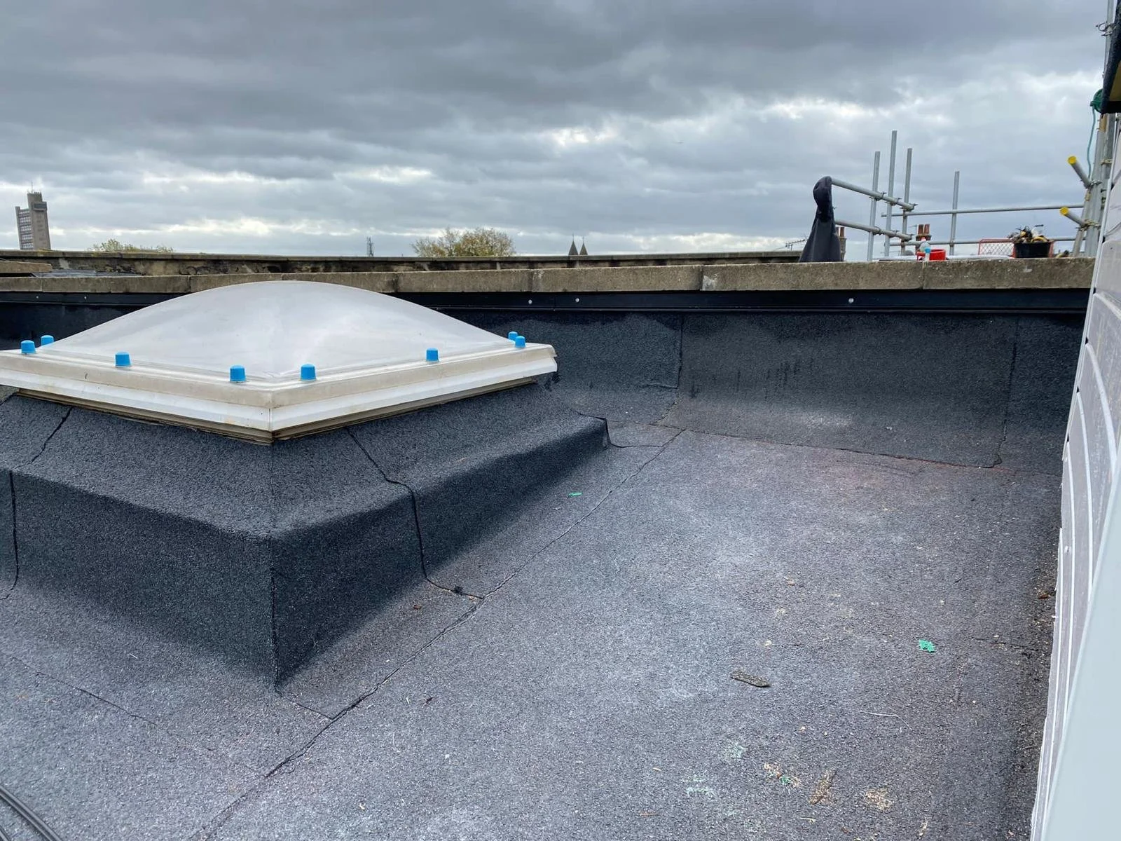 A rooftop with a domed skylight, asphalt roofing material, and some construction scaffolding in the background under a cloudy sky.