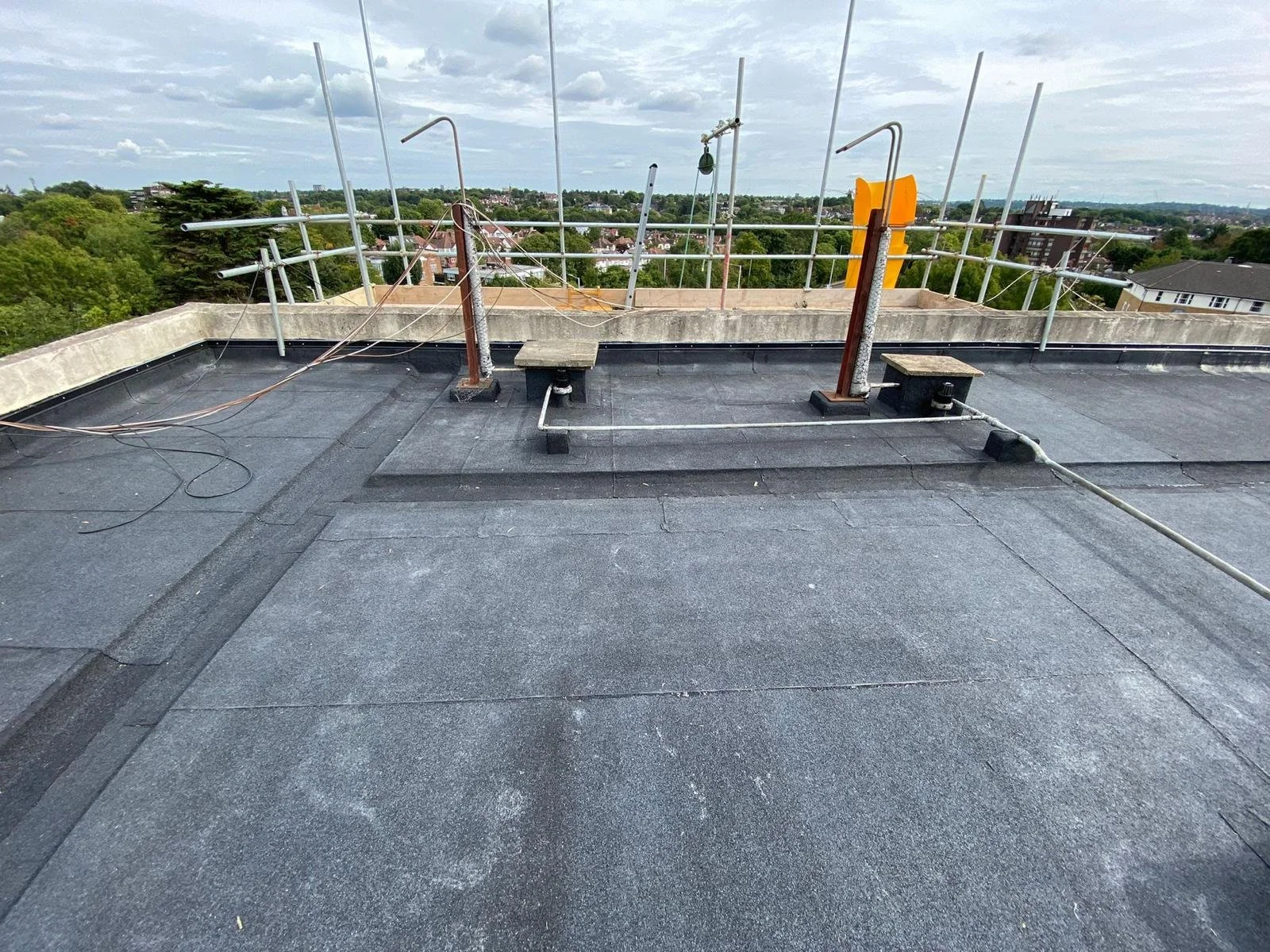 View of a rooftop with black roofing material, metal railings, and height antennas, overlooking a suburban landscape with trees and houses under a cloudy sky.
