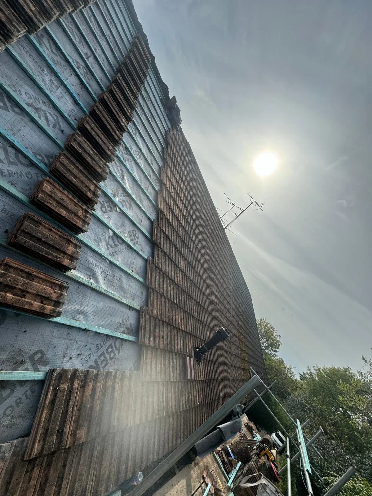 Building under construction with scaffolding and brickwork, under a cloudy sky with the sun visible, roofing materials, and construction equipment.