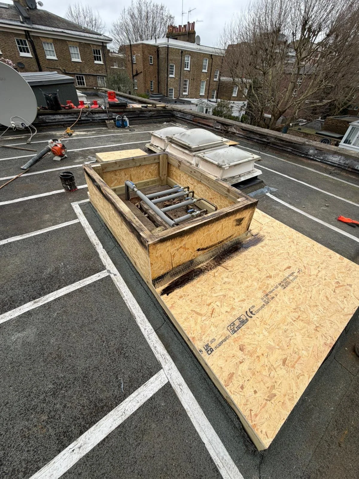 A rooftop under construction with a wooden frame and pipes, surrounded by tools and building materials, in an urban area with brick buildings in the background.