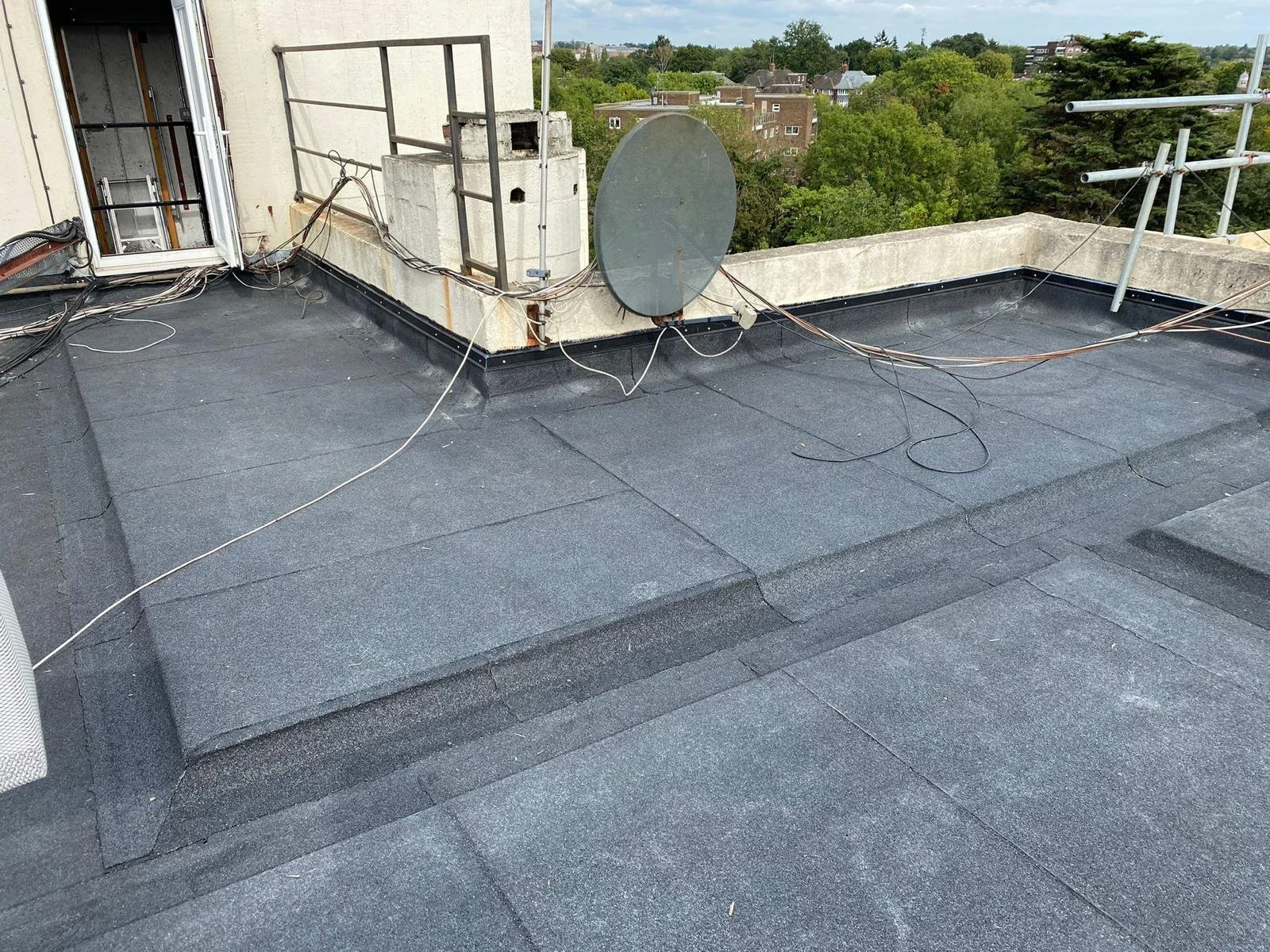 The rooftop has black asphalt shingles, with various wires and cables, a satellite dish, and scaffolding, overlooking a green residential area with trees and buildings in the background.