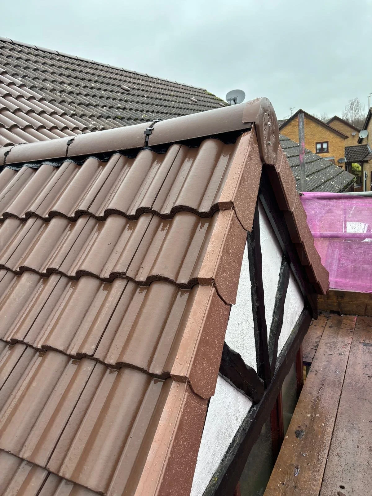 Close-up view of a sloped roof with brown ceramic tiles and black trim, showing waterproofing details, with a background of neighboring houses and an overcast sky.