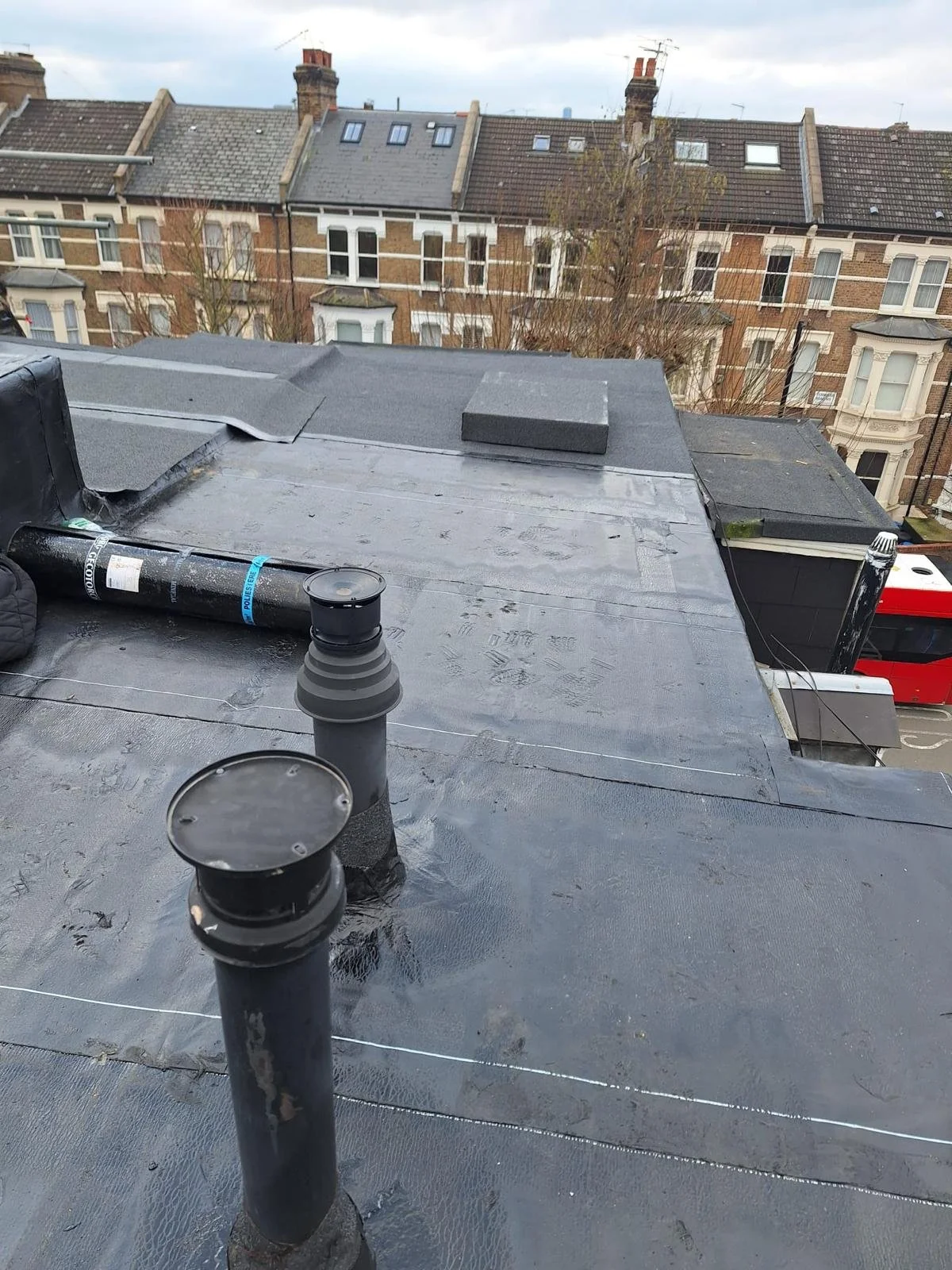 Flat rooftop with vents, chimney, and black waterproof coating, overlooking neighboring buildings and cloudy sky.