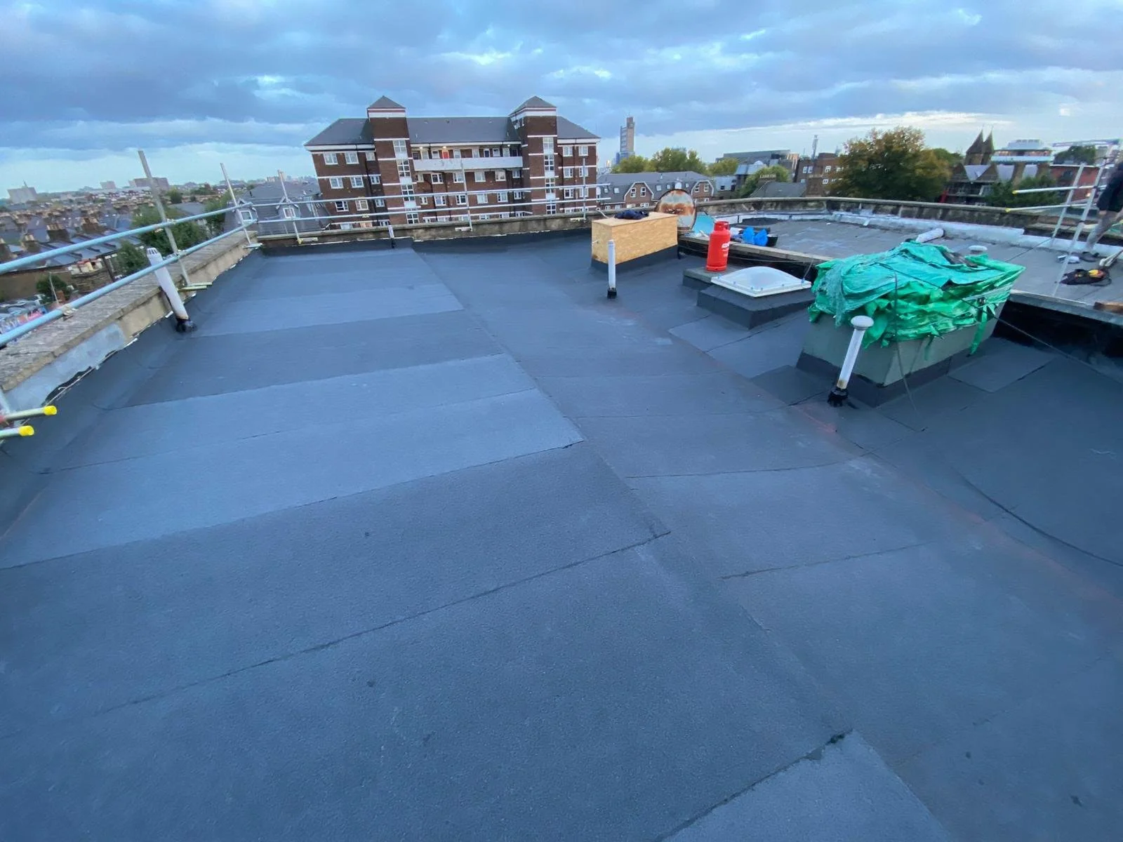 A rooftop with black waterproofing material, safety railing, and various equipment and materials for maintenance or repairs, with cityscape and cloudy sky in the background.