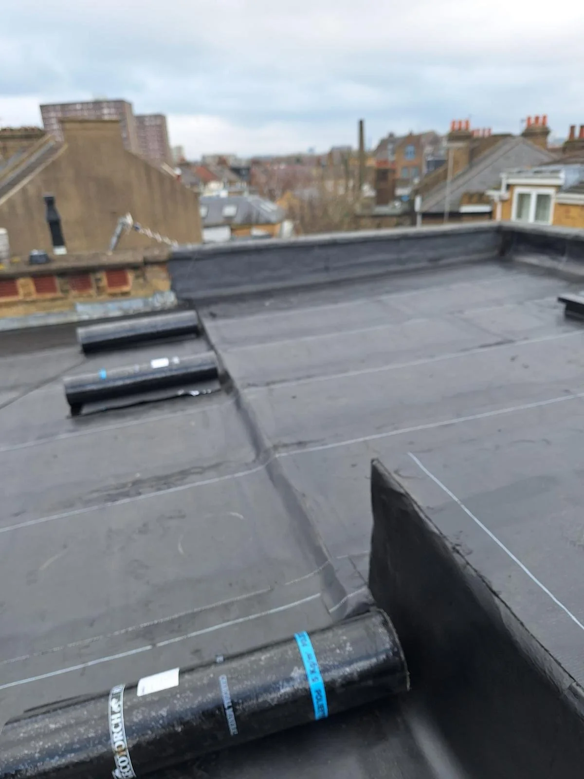 Flat rooftop with black roofing material, ventilation vents, and cityscape in the background under cloudy sky.