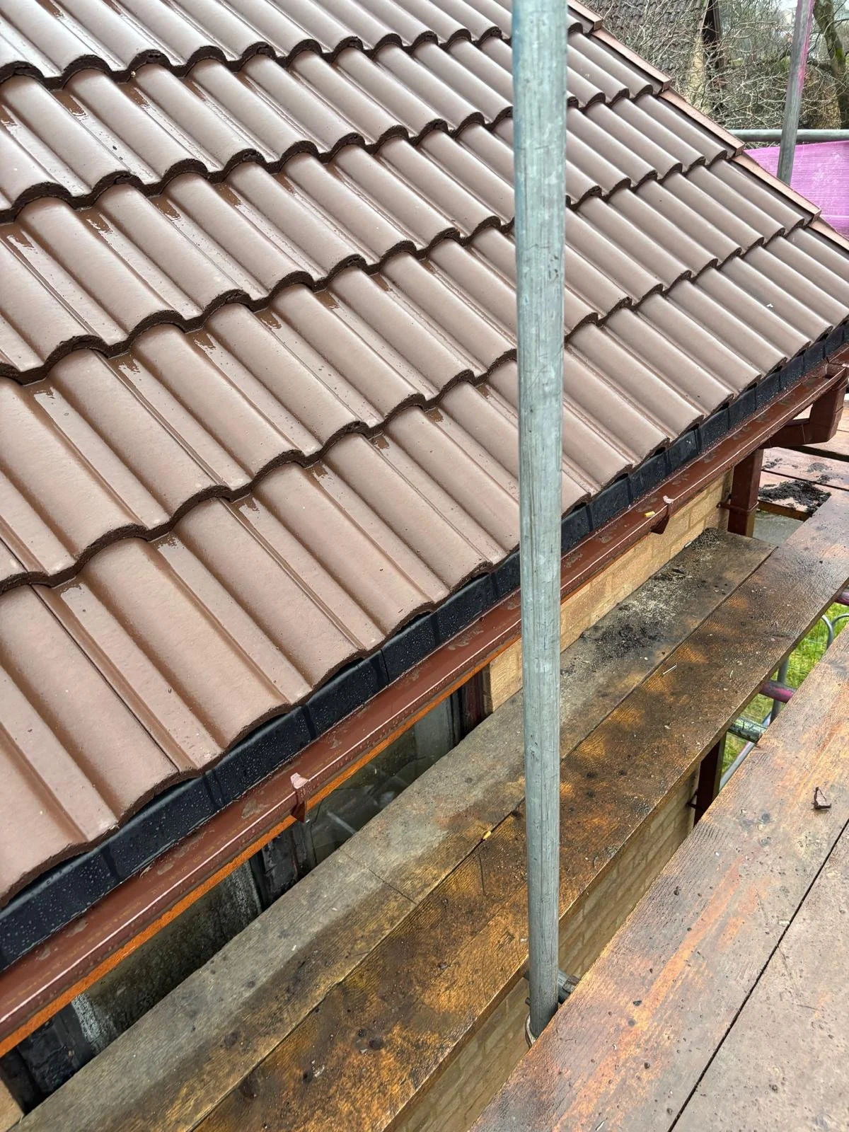 Close-up of a house roof with new brown tiles, with scaffolding and wooden planks nearby, indicating ongoing construction or renovation.