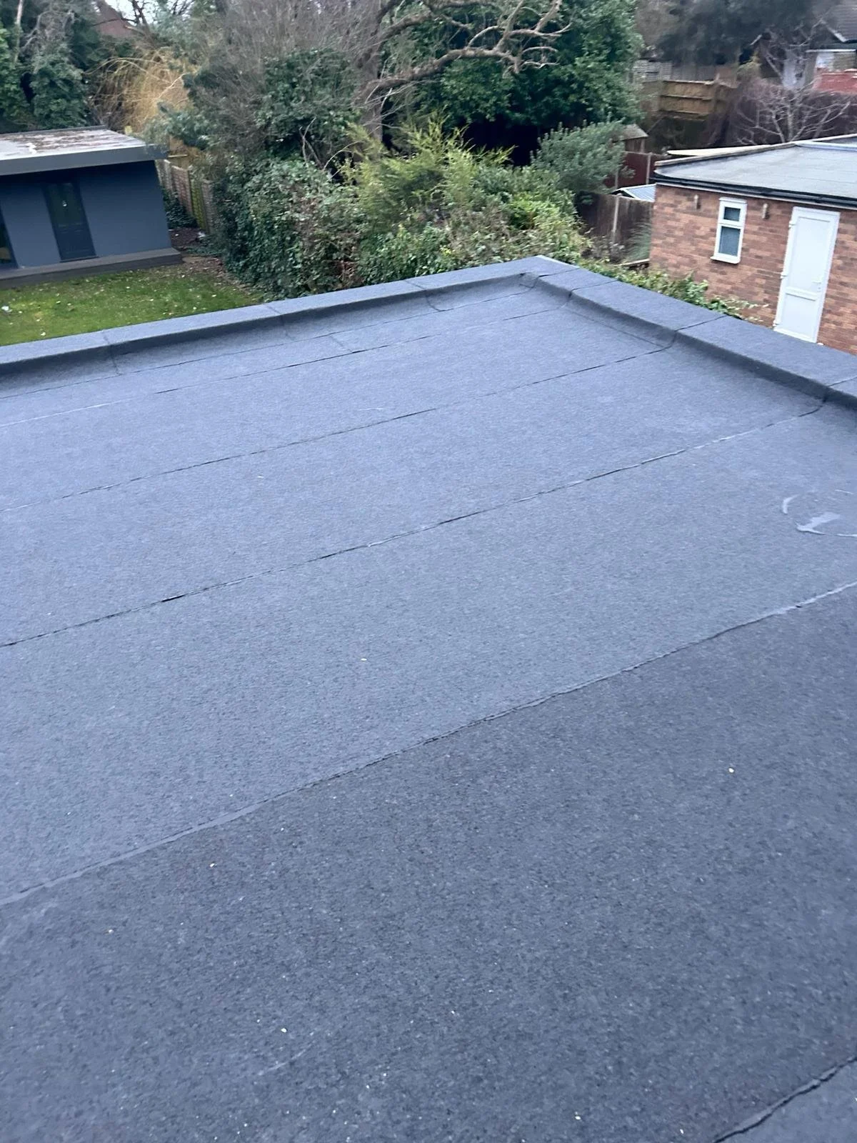 View of a flat roof covered with dark gray asphalt shingles, with trees and neighboring houses in the background.