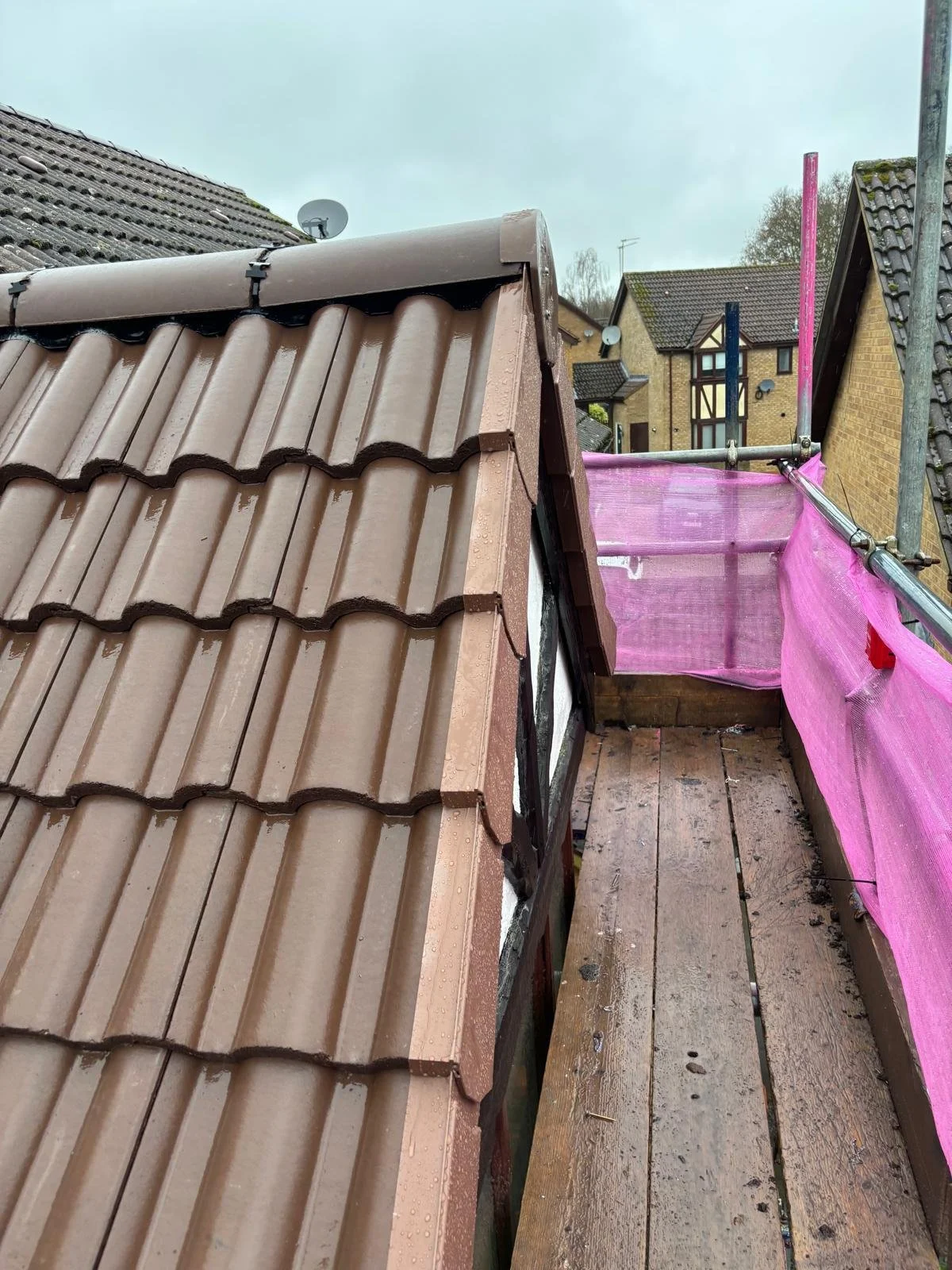 Roof construction site showing new brown tile roofing, scaffolding with pink safety netting, and wooden platform with surrounding residential houses.
