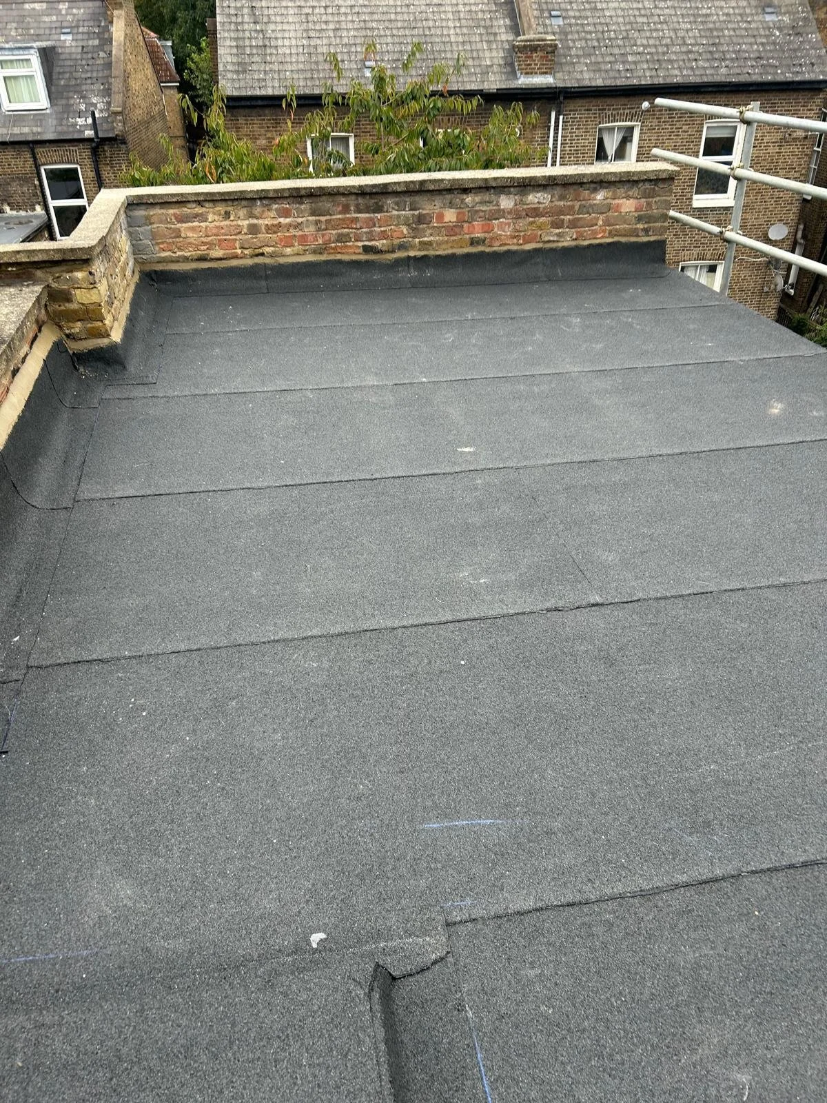 Flat rooftop covered with black roofing material, brick wall with some greenery in the background, and neighboring brick buildings with chimneys and windows.