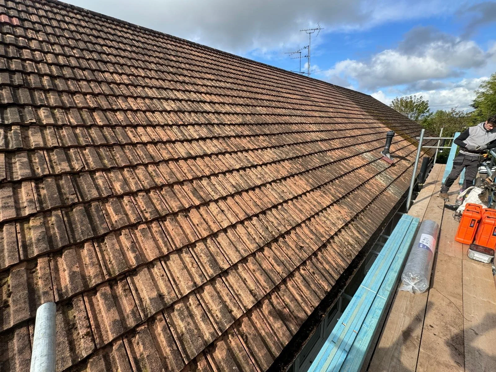 View of a roof with weathered red-brown tiles, construction materials, and workers on a scaffolding platform.