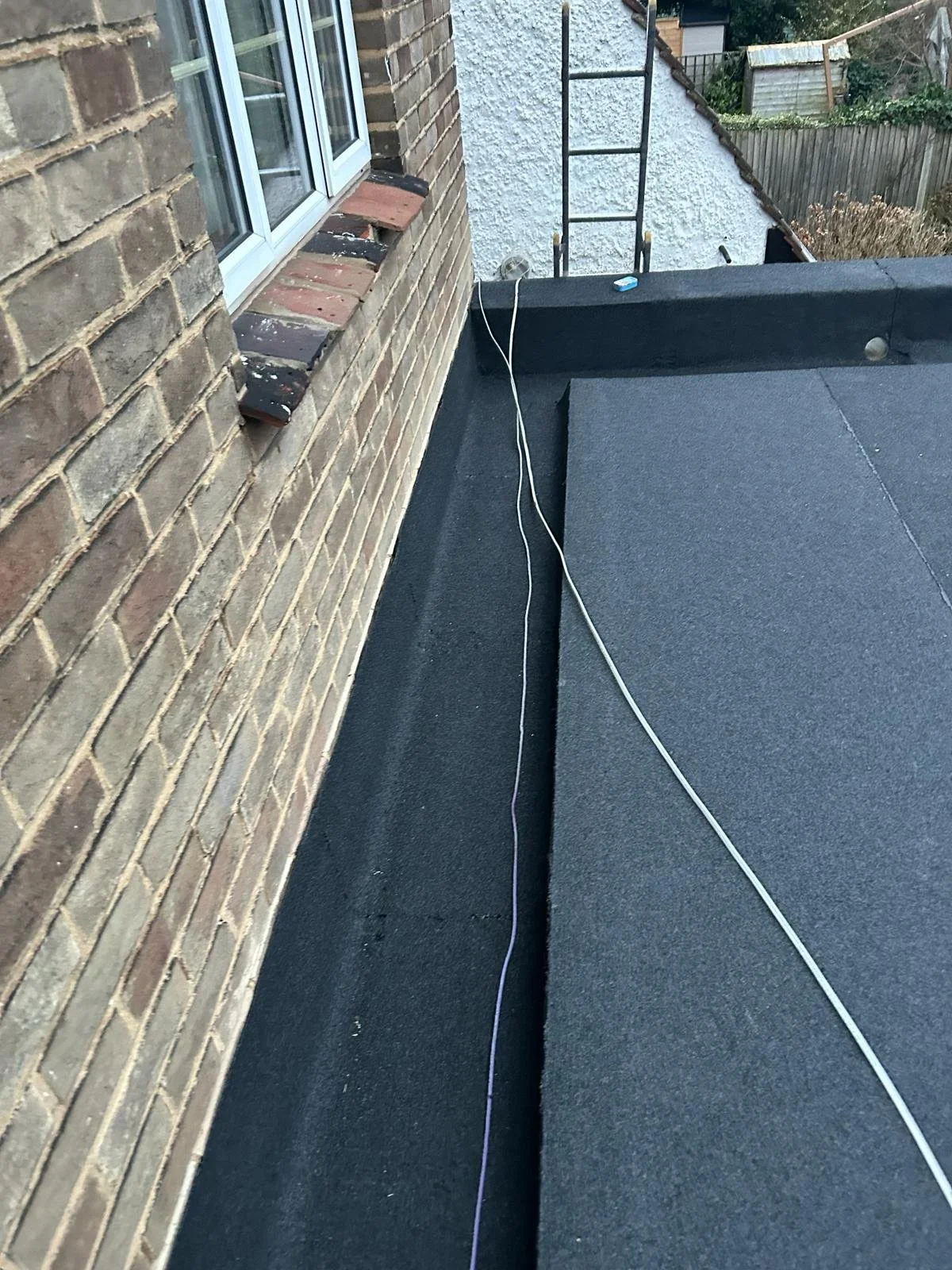 The roof of a building with a black surface, a brick wall with a window, a ladder, and a string or wire stretched across the roof. In the background, there are trees and a neighboring building with a sloped roof.