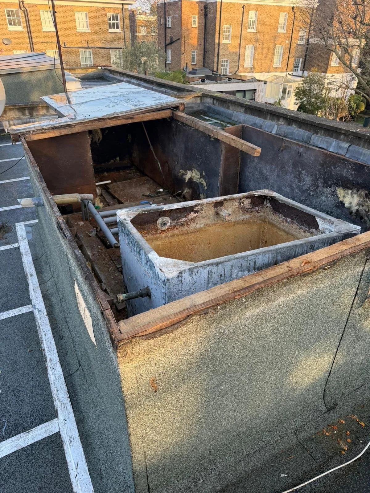 Rooftop HVAC unit with rooftop access hatch removed, exposing internal components and piping, with surrounding urban buildings and parking in background.